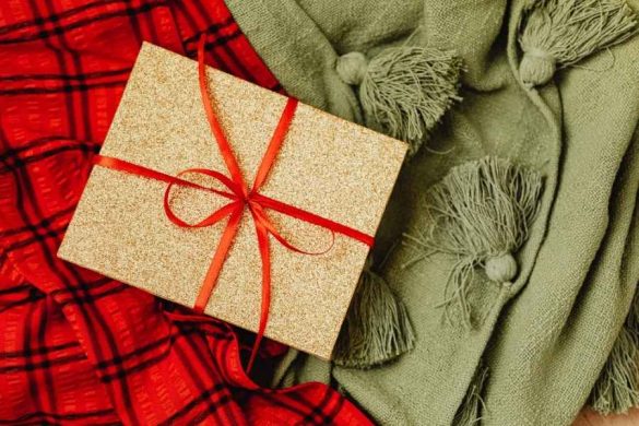Coffret cadeau doré avec ruban rouge posé sur une écharpe verte parmi des cadeaux originaux