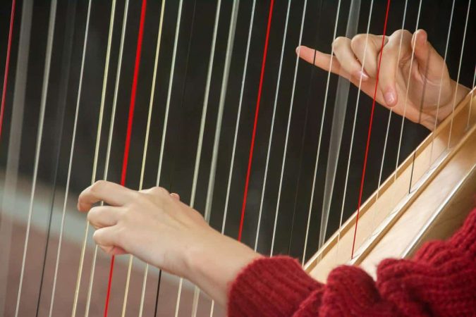 Gros plan sur des mains jouant les cordes d'une harpe avec un pull rouge, illustrant l’apprentissage de la harpe.