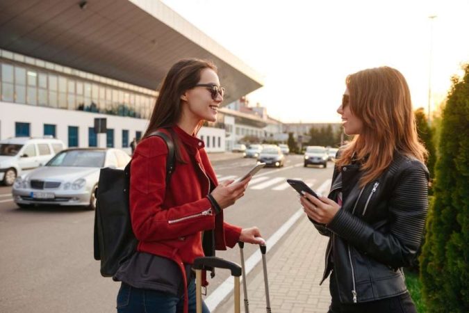 Deux amies discutent avec leur smartphone devant un terminal d'aéroport, prêtes pour leurs déplacements à l'étranger.