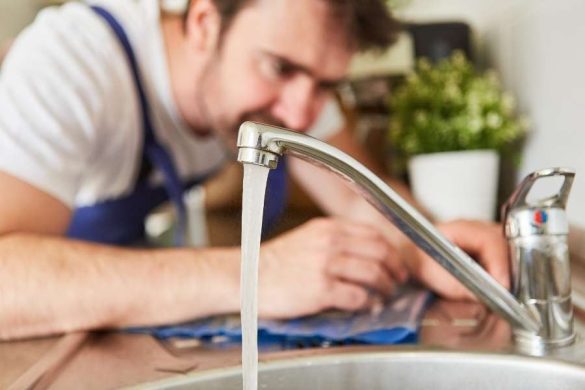 Un plombier concentré inspecte et répare un vieux robinet mitigeur de cuisine chromé dont l'eau coule dans l'évier en inox.